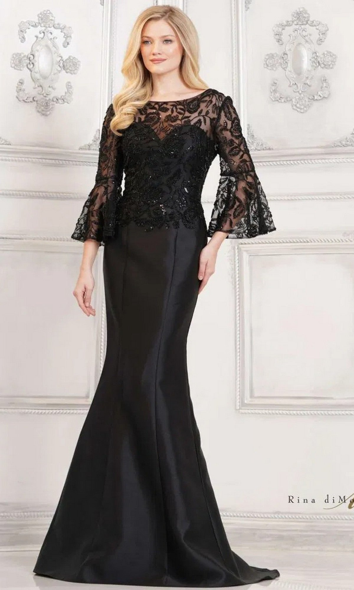 A woman in a black Rina Di Montella RD2939 long formal dress with lace bell sleeves, a beaded bodice, and a fitted mermaid skirt stands against an ornate white wall.
